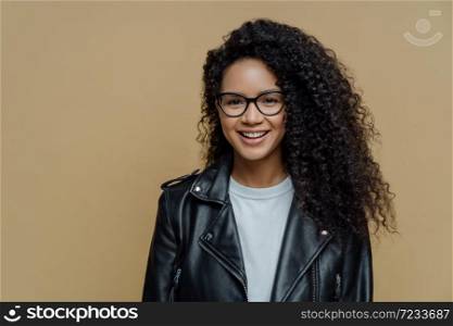 Stylish hipster girl with bushy Afro haircut, smiles positively, enjoys nice day, wears spctacles and black leather jacket, poses against brown background. People, emotions and fashion concept