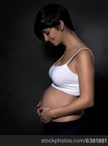Stylish Hispanic pregnant woman standing side view over black background, looking on her tummy and touching it, healthy pregnancy, enjoying motherhood