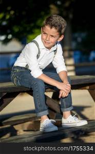 Stylish little boy in white shirt and suspenders posing in park and looking away.. Dandy kid posing in sunlight
