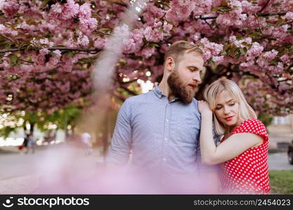 Stylish lovers near the blossoming Sakura. beautiful young couple, man with beard and blonde woman hugging and having fun in the spring park. Concept spring. fashion and beauty.. Stylish lovers near the blossoming Sakura. beautiful young couple, man with beard and blonde woman hugging and having fun in the spring park. Concept spring. fashion and beauty