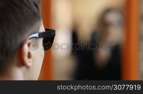 Stylish man looking for the best sunglasses in optical store. Handsome young man reflected in big mirror trying on new sunglasses in optician shop. Selective focus.