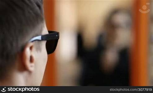 Stylish man looking for the best sunglasses in optical store. Handsome young man reflected in big mirror trying on new sunglasses in optician shop. Selective focus.
