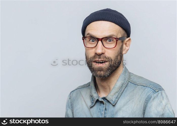 Stylish middle aged male with suprised and curious expression wears spectacles, denim jacket and hat, poses against grey background with copy space for your advertisment or promotional text.