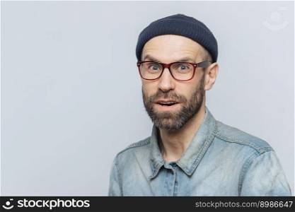 Stylish middle aged male with suprised and curious expression wears spectacles, denim jacket and hat, poses against grey background with copy space for your advertisment or promotional text.