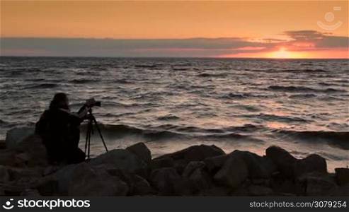 Stylish professional photographer with backpack making photos of amazing red sunset and seascape with camera on a tripod in twilight time. Skillful man photographing landscape and sea in glow of amazing setting sun on the beach. Slow motion.