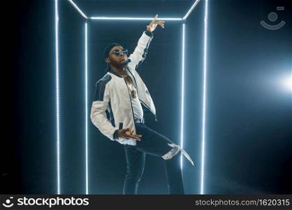 Stylish rapper in sunglasses poses in illuminated cube, dark background. Hip-hop performer, rap singer, break-dance performing, entertainment lifestyle. Rapper in sunglasses poses in illuminated cube