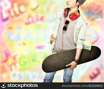 Stylish teen boy with skateboard in hand and earphones on the neck over colorful background, active guy has a cool hobby, modern life of young people