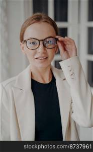 Stylish young caucasian woman fixes glasses on face, looking at camera. Pretty smiling businesswoman wearing trendy eyewear posing for business portrait. Advertisement of eyeglass store.. Stylish female fixes trendy glasses, posing for business portrait. Advertisement of eyeglass store