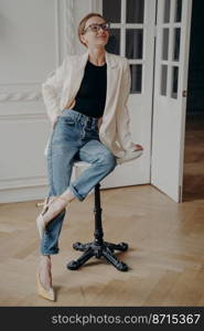 Stylish young female wearing casual clothes, sitting on little round table indoors. Smiling businesswoman fashion model in glasses, blue jeans and white suit jacket posing in interior indoors.. Stylish businesswoman in glasses wearing casual clothes posing sitting on little round table indoors