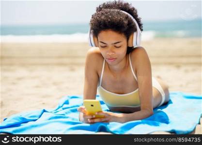 Stylish young fit African American female millennial with dark curly hair, in trendy bikini and headphones listening to music, and messaging on smartphone while lying on sandy seashore in sunlight. Young ethnic female tourist using smartphone and listening to music on beach