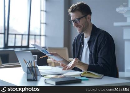 Stylish young German businessman in glasses discussing calculation chart reports with partners online via video call on laptop while sitting at desk at home office. Happy freelancer having web meeting. Young German businessman in glasses discussing calculation chart reports with partners online