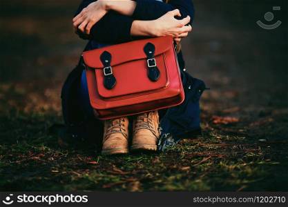 stylish young girl in brown shoes and warm coat sits in the park with a red bag.. stylish young girl in brown shoes and warm coat sits in the park with a red bag