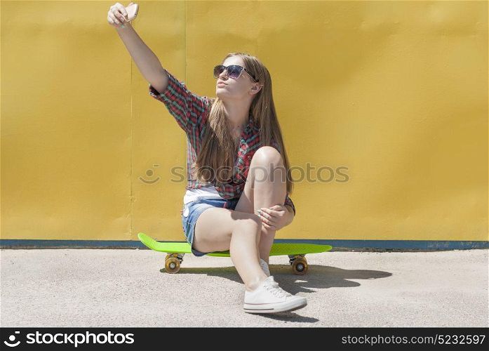 Stylish young girl sitting on a skateboard makes a self-portrait smartphone.