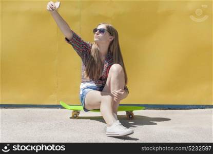 Stylish young girl sitting on a skateboard makes a self-portrait smartphone.