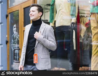 stylish young man wearing jacket with sa≤tag standing outside shop