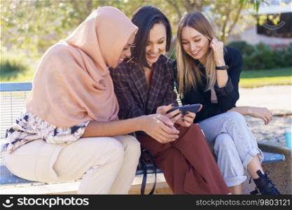 Stylish young multiracial female friends smiling while watching interesting video on mobile phone sitting on bench in park on sunny day. Smiling young diverse women watching video on smartphone in park
