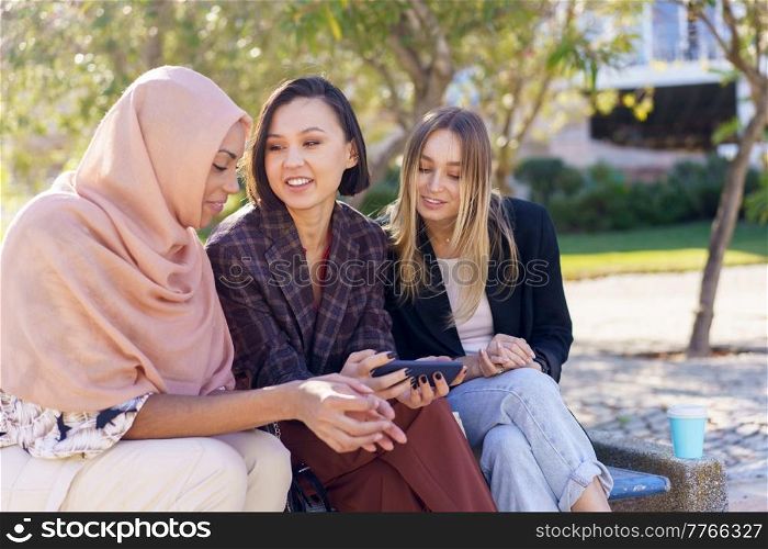 Stylish young multiracial women friends, smiling while watching interesting video on mobile phone sitting on bench in park on sunny day. Smiling young diverse girls watching video on smartphone in park