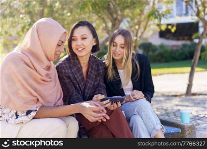 Stylish young multiracial women friends, smiling while watching interesting video on mobile phone sitting on bench in park on sunny day. Smiling young diverse girls watching video on smartphone in park