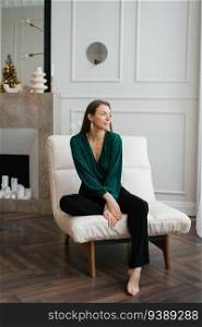 Stylish young woman fashion blogger in an emerald blouse and black trousers sits on a white modern armchair in the living room and looks away