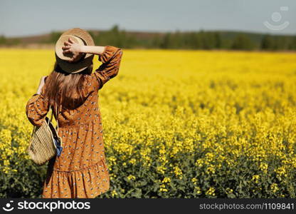 stylish young woman in a field of yellow flowers. Girl in straw hat and in a floral dress and with a wicker bag. place for inscription. rear view.