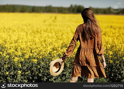 stylish young woman in a field of yellow flowers. Girl in straw hat and in a floral dress. place for inscription. rear view. background with yellow flowers and blue sky.