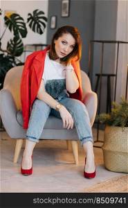 stylish young woman in white t shirt, jeans and red jacket and shoes on high heels is sitting on chair and looking in camera. fashion concept. studio short.. stylish young woman in white t shirt, jeans and red jacket and shoes on high heels is sitting on chair and looking in camera. fashion concept. studio short
