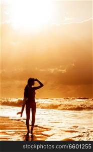 Stylized saturated rear view of beautiful sexy young woman surfer girl in bikini with white surfboard on a beach at sunset or sunrise