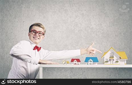 Suburban life. Young man in glasses pointing at house model