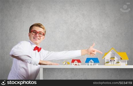 Suburban life. Young man in glasses pointing at house model
