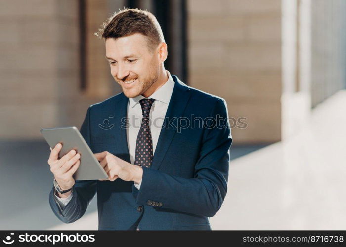 Succesful male entrepreneur in formal suit, holds modern tablet, checks documentation online, reads necessary information for making business report, uses free wifi connection. People and technology