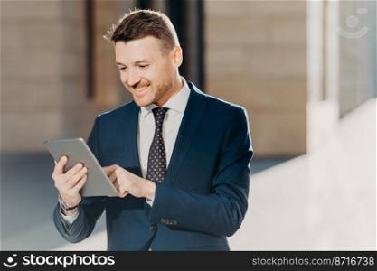Succesful male entrepreneur in formal suit, holds modern tablet, checks documentation online, reads necessary information for making business report, uses free wifi connection. People and technology