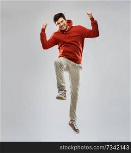 success, achievement and motion and people concept - smiling young man in red hoodie jumping over grey background and celebrating victory. man in hoodie jumping and celebrating success