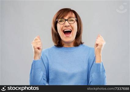 success and old people concept - portrait of happy laughing senior woman in glasses celebrating triumph over grey background. portrait of happy senior woman celebrating triumph