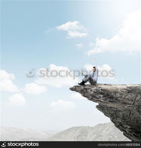 Success in business. Young businessman sitting on edge of rock mountain and using tablet pc