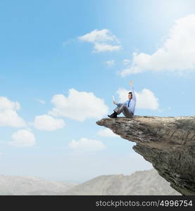 Success in business. Young businessman sitting on edge of rock mountain and using tablet pc