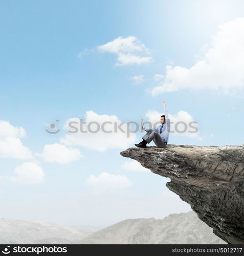 Success in business. Young businessman sitting on edge of rock mountain and using tablet pc