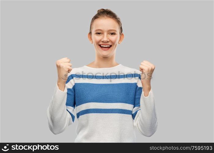 success, triumph and people concept - happy smiling teenage girl in pullover celebrating victory over grey background. happy teenage girl in pullover celebrating success