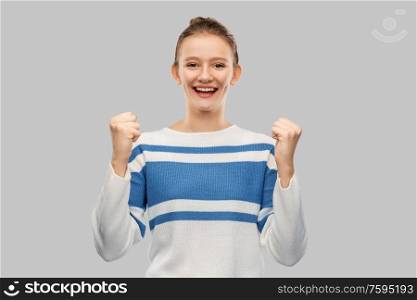 success, triumph and people concept - happy smiling teenage girl in pullover celebrating victory over grey background. happy teenage girl in pullover celebrating success