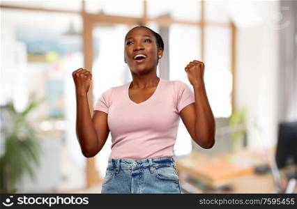 success, winning gesture and people concept - happy smiling young african american woman celebrating victory over office background. happy african american woman celebrating success