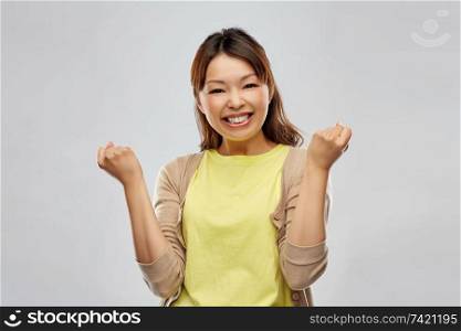 success, winning gesture and people concept - happy young asian woman celebrating victory over grey background. happy asian woman celebrating success