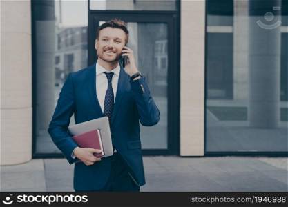 Successful and confident business owner in formal wear, talking on phone with his employee, telling directions and instructions on next move, holding note book with laptop, blurred background. Successful and confident business owner talking on phone with his employee outdoors
