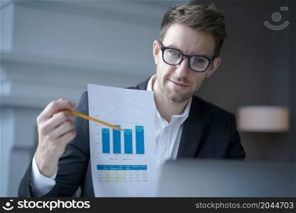 Successful Austrian CEO male in formal suit pointing at charts and interest rate in pencil while sitting in front of laptop at home office interior, communicating with new business partners online. Austrian CEO male in suit sitting in front of laptop at home and explaining financial report online