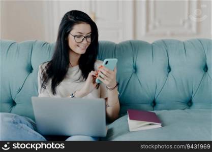 Successful brunette freelancer watches live stream, poses with gadgets on sofa, reads cellphone messages, works casually from home.