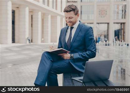 Successful business owner in formal wear writing down information he is receiving from conference call on laptop, sitting on bench with crossed legs and working remotely, doing freelance job outdoors. Successful business owner writing down information in notebook while working outdoors on weekend