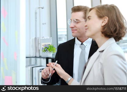 Successful businessman and businesswoman writing good ideas on sticky notes pasted on glass wall in modern office meeting.