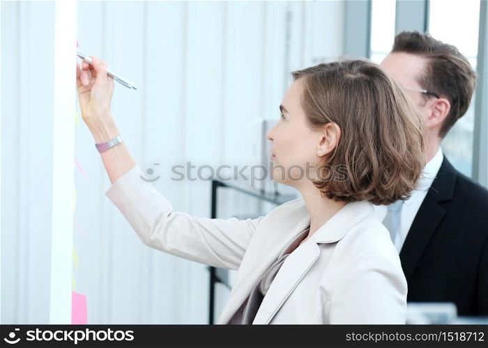 Successful businessman and businesswoman writing good ideas on sticky notes pasted on glass wall in modern office meeting.