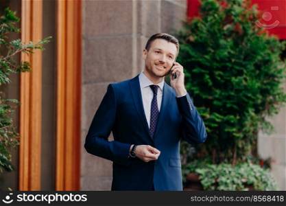 Successful businessman has work break, uses mobile phone for having talk, being always on line, dressed in formal suit, has pleasant smile on face, stands outdoor near restaurant. Technology concept
