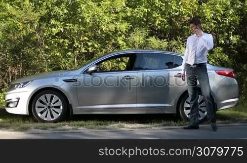 Successful businessman in formal wear discussing big deal agreement with foreign partner using smartphone near his car. Emotional business executive arguing with colleague on mobile phone while walking in front of parked vehicle on rural roadside.