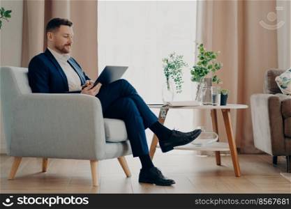 Successful businessman in formal wear sits in comfortable armchair uses modern laptop prepares online project poses against home interior analyzes financial indicators controls company development