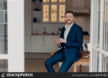 Successful businessman in suit typing message on mobile phone or reading news and smiling at camera while leaning on wooden table in living room of his modern apartment. Business people concept. Bearded guy entrepreneur in suit using modern gadgets in his apartment